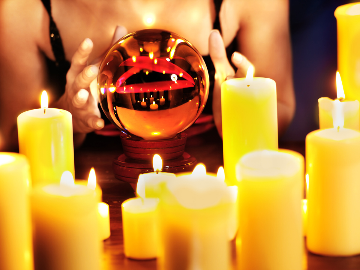 A Psychic woman’s hands are around a crystal ball with several burning candles in the foreground.